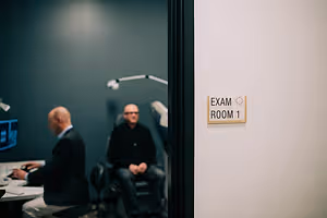 Two men sit in an exam room, one of them appears to be waiting for an appointment.