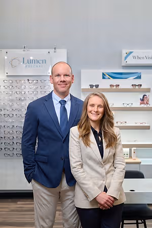 A man and a woman standing together in an office room with a table, chairs, and shelves with glasses, and a sign that reads 'Lumen Eyecare'.