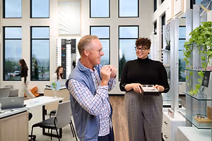 A man and a woman are standing inside a room with wooden floors and large windows. The man is wearing glasses and a blue vest, while the woman is holding a box and wearing glasses and a black turtleneck. There are several people walking in the distance, and there are chairs, a table with a laptop, a plant, and a shelf with a plant in the room.