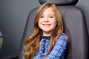 A young girl with long blonde hair and blue checkered shirt is smiling while sitting on a black chair with leather upholstery in what seems to be a dental clinic.