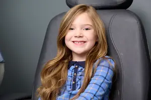 A young girl with long blonde hair and blue checkered shirt is smiling while sitting on a black chair with leather upholstery in what seems to be a dental clinic.