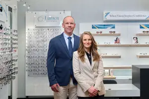 A man and a woman stand side by side in front of a wall with various eyeglass displays and a sign that reads When Vision Inspires Confidence