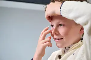 A young boy with a light blue sweater puts a contact lens in his eye.