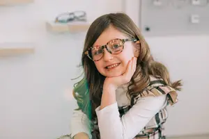 A girl wearing glasses is sitting and smiling while posing for a photo.