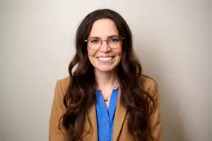 A woman with brown hair wearing glasses, a necklace, a blue shirt, and a brown blazer is smiling and posing for a photo in front of a white wall.