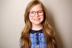 A young girl with long blonde hair wearing red-framed glasses and a blue checkered shirt is smiling and seems to be posing for a photo against a light-colored wall.