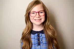 A young girl with long blonde hair wearing red-framed glasses and a blue checkered shirt is smiling and seems to be posing for a photo against a light-colored wall.