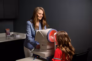 A woman in a blue blazer is helping a girl with long hair use a machine to check her eyes in a room with a black wall and a white countertop with a book on it.