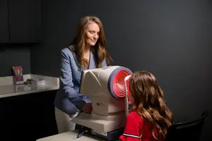 A woman in a blue blazer is helping a girl with long hair use a machine to check her eyes in a room with a black wall and a white countertop with a book on it.