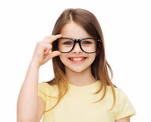 A young girl in a yellow t-shirt wearing glasses and smiling with one hand on her forehead