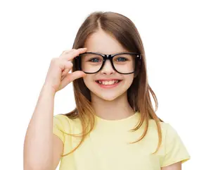 A young girl in a yellow t-shirt wearing glasses and smiling with one hand on her forehead