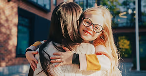 A young girl with glasses and a yellow and white sweater is hugging a girl with long hair. They are smiling and posing for a photo in front of a building.