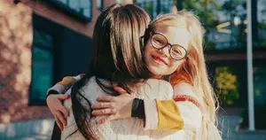 A young girl with glasses and a yellow and white sweater is hugging a girl with long hair. They are smiling and posing for a photo in front of a building.