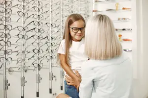 An adult woman and a young girl are standing in a store with rows of eyeglasses on display. They are looking at each other with smiles on their faces.