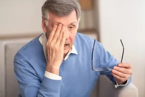 An old man with glasses covering his eyes while holding them in his right hand.
