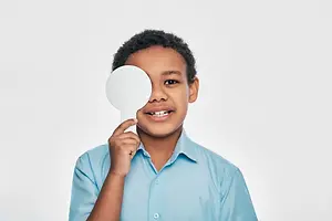 A young boy is holding a mirror in front of his eye and smiling at the camera.