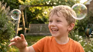 A young boy is smiling and holding a bubble wand while blowing bubbles in a garden