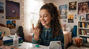 A girl is sitting at a desk in her bedroom, smiling and making a peace sign with her hand. There are various items on the desk, including a book, a cup with pencils, a bottle, and a small box. Behind her, there is a bed with a pink blanket and a shelf with books. Posters are on the walls, and a skateboard is leaning against the wall.