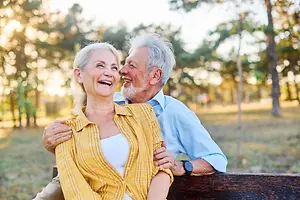 An elderly couple sitting on a bench, both smiling and looking at each other in a park