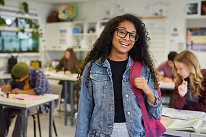 A girl with glasses and a denim jacket stands in a classroom with other students, one of whom is writing on a piece of paper