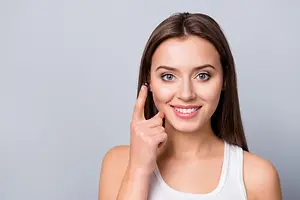 A woman with long hair wearing a white sleeveless top is smiling and posing for a photo with a finger on her lips and a finger pointing at her eye.