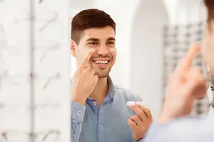 A man is trying on contact lenses in front of a mirror, with a blurry person in the background.