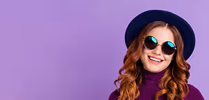 A woman in a purple turtleneck and sunglasses smiling standing against a purple background