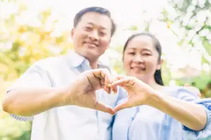 An older couple with gray hair smiles and forms a heart shape with their hands