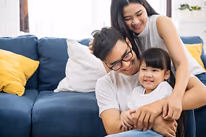 A family sitting on a couch, with a man wearing glasses and a woman wearing a sleeveless top holding a child, all smiling at the camera.
