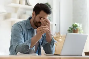Man in denim shirt sitting at a table, holding glasses and rubbing his eyes, with a laptop in front of him.