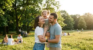 A family with a young child is smiling in a park, surrounded by others enjoying the outdoors.