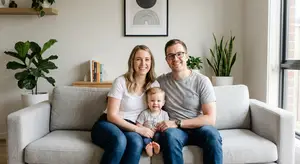 A family of three people, a woman, a man, and a child, are sitting on a gray couch with a smile on their faces. They are in a room with a gray wall, a wooden shelf, and several potted plants.