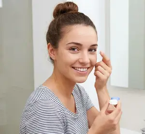 A woman smiles in the mirror while inserting a contact lens into her eye.