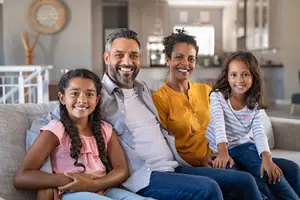 Four members of a family sitting together on a couch, two young girls and their parents, all smiling at the camera.