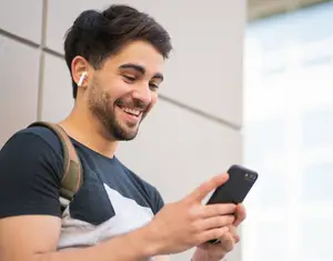 A young man wearing a backpack is smiling and looking at his smartphone while standing in front of a building. He has a pair of white earphones in his ears.
