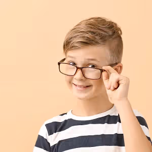 A boy with brown hair wearing a striped shirt and eyeglasses is smiling and posing for a picture with his left hand on his face.