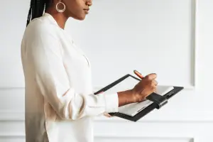 A woman is writing on a notepad while standing in front of a white wall
