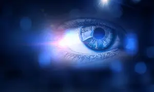 Close up of a human eye with a blue iris and a white pupil, surrounded by eyelashes, with a blue light shining on it, against a dark blue background