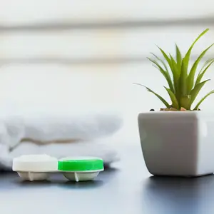 Two green and white lens cases sit next to a plant in a white pot on a table with towels in the background.