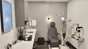 A medical examination room featuring an optometrist's chair, a wall-mounted monitor, a desk with a computer monitor and keyboard, and a trash bin.
