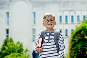 A smiling boy with glasses and a backpack holding a book in front of a building