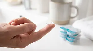 A hand holding a contact lens above a table with a cup, towels, and contact lens packages