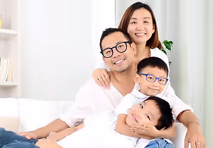 A family sitting on a white couch with two kids wearing glasses and the mother with her hand on the man's shoulder