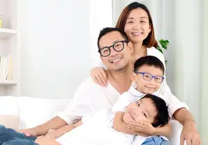 A family sitting on a white couch with two kids wearing glasses and the mother with her hand on the man's shoulder