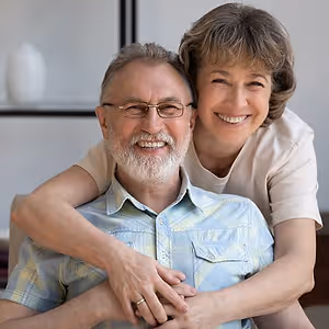 An elderly man wearing glasses and a woman with short hair are smiling and posing for a photo with their arms around each other