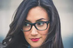 A woman with long black hair wearing glasses and a blue shirt is smiling and looking to the left. She has a mole on her left cheek and is standing in front of a white wall.