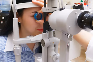 woman with long black hair wearing a white uniform with blue floral print and a silver earring getting her eye examined with a medical ophthalmoscope