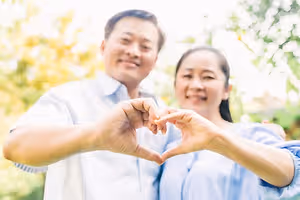A couple is posing for a picture with their hands shaped like a heart, smiling and standing near a tree in a park during the daytime