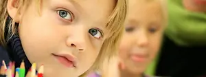 Two children, one with blonde hair and blue eyes and the other with blonde hair, are sitting next to each other in a classroom. The child with blonde hair and blue eyes is holding a pencil and appears to be looking at something. Behind her, another child is looking to the right. Both children are wearing black and purple tops. There are several pencils on the left side of the image.
