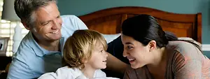 A man, woman, and child are lying on a bed and smiling at the camera.